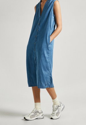 Vrouw draagt een mouwloze blauwe midi-jurk met knopen aan de voorkant en zakken, crèmekleurige sokken en witte en grijze sneakers, staand tegen een effen achtergrond.