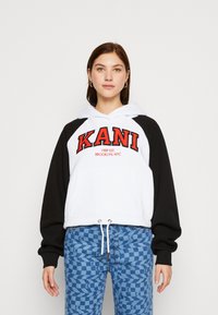 Hanorac cu glugă, cu corp alb și mâneci negre, având un mare logo roșu "KANI" și textul "1989 EST. BROOKLYN, NYC". Snur la tiv.