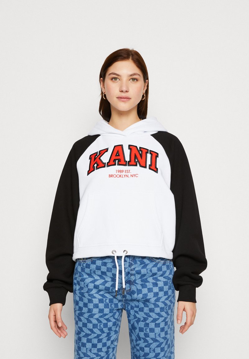 Hanorac cu glugă, cu corp alb și mâneci negre, având un mare logo roșu "KANI" și textul "1989 EST. BROOKLYN, NYC". Snur la tiv.