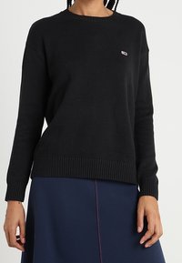 Pull noir en maille avec un col rond et des poignets côtelés. Présente un petit écusson logo sur la poitrine. Coupe ample avec une surface texturée.