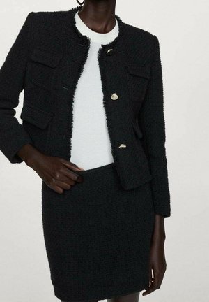 Vrouw draagt een zwart gestructureerd jasje met knopen en voorzakken over een wit topje en een bijpassende zwarte rok.