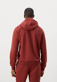 Muž viděný zezadu, oblečený v vínovém mikině s kapucí a odpovídajících teplácích, proti jednoduchému bílému pozadí.