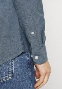 Modrá košile chambray s dlouhými rukávy, manžetami na dvojité knoflíky a zakřiveným lemem. Nositelná přes vybledlé džíny s viditelnými švovými detaily.