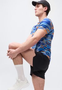 Νέος άνδρας με μπλε πουκάμισο με σχέδια, μαύρο σορτς, λευκά αθλητικά παπούτσια και μαύρο καπέλο κρατά το γόνατό του στο στήθος σε μια στάση διάτασης μπροστά σε λευκό φόντο.