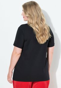 Černé tričko s krátkým rukávem a volným střihem, vyrobené z měkkého materiálu, v kombinaci s červenými skládacími kalhotami. Viděno zezadu.