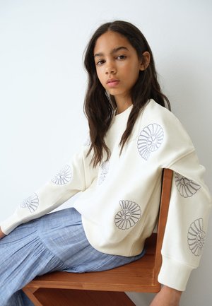 Jeune femme aux longs cheveux foncés assise sur une chaise en bois, portant un pull crème avec des motifs de coquillages en spirale et une jupe plissée bleu clair.