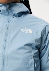 Jasnoniebieska wodoodporna kurtka z kapturem, wyposażona w przedni zamek błyskawiczny oraz biały logo "The North Face" na górnej lewej stronie.