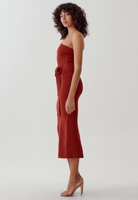 Vestido rojo ceñido con diseño sin tirantes y un detalle de lazo en la cintura. La tela tiene una textura suave y un corte por la rodilla.
