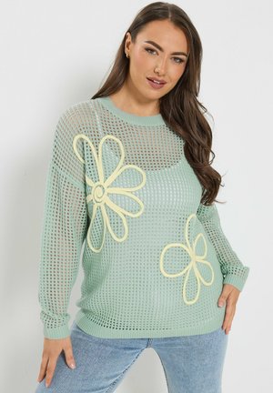 Frau trägt einen offen gestrickten mintgrünen Pullover mit großen gelben Blumenmustern, kombiniert mit hellblauen Jeans, und steht vor einer weißen Wand.