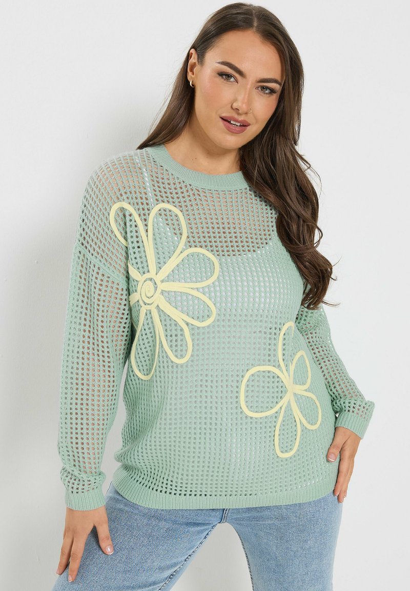 Kvinde iført en mintgrøn åbenstrikket sweater med store gule blomstermotiver, kombineret med lyseblå jeans, stående op ad en hvid væg.