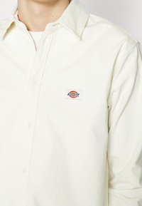 Camisa blanca de pana con diseño abotonado, con una pequeña etiqueta "Dickies" en rojo, amarillo y azul en el área izquierda del pecho.