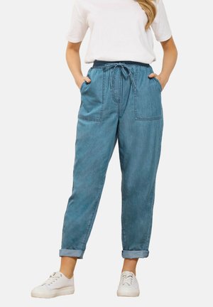 Kvinde iført lyseblå denim joggerbukser med opsmøgede kanter, elastisk talje med snøre, forlommer, hvide sneakers og hvid T-shirt.