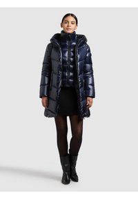 Tamsiai mėlynas pūstas paltas su gobtuvu, blizgiu paviršiumi ir siūtu dizainu. Dėvimas ant derančios trumpintos striukės ir ribuoto sijono.