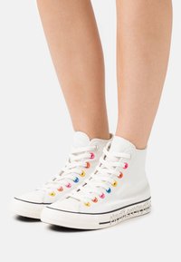 Scarpe alte in tela bianca con occhielli colorati, suola in gomma e un distintivo logo bianco sul lato. Allacciate con lacci bianchi.