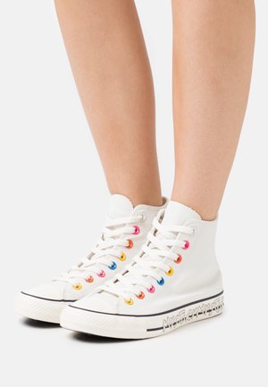Baskets montantes en toile blanche avec des œillets colorés, semelle en caoutchouc et logo blanc distinctif sur le côté. Lacées avec des lacets blancs.