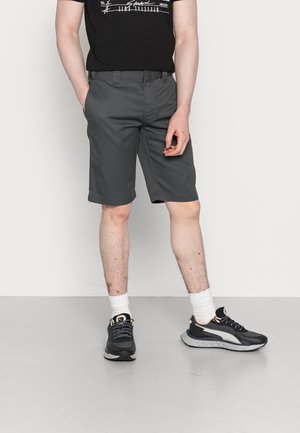 Personne portant un short gris foncé jusqu'aux genoux, des chaussettes blanches et des baskets noires, debout devant un fond clair uni.