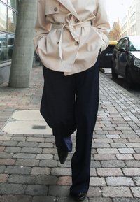 Beež kahekordne vihmajope, millel on sidumisrihm, lahtiste tumedate siniste pükste peal, mustade teravikute jalatsitega. Taustal kõnnitee kividega.