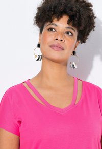 Fuchsie krátký rukáv top s výstřihem do V a dvojitými ramínky. Zlaté kruhové náušnice mají černé a bílé kruhové akcenty.