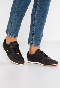 Svarta träningsskor med en slät tygöverdela, snören, guldiga accenter och en vit gummisula. Mönstrade detaljer vid hälen och sidorna.