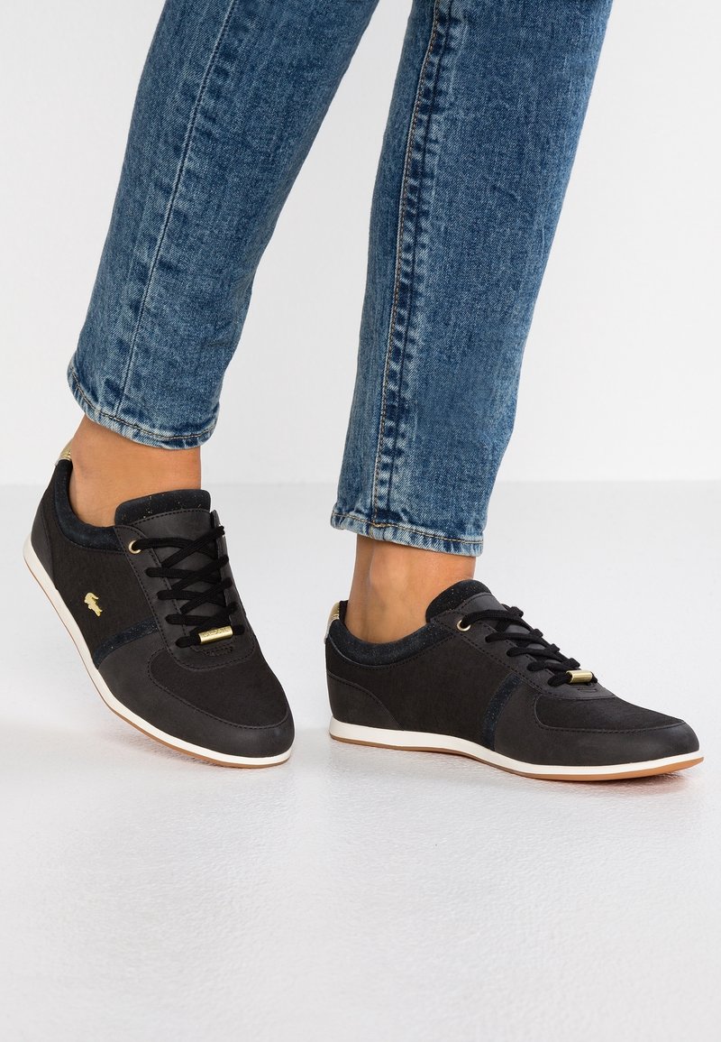 Svarta träningsskor med en slät tygöverdela, snören, guldiga accenter och en vit gummisula. Mönstrade detaljer vid hälen och sidorna.