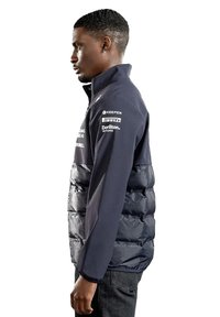 Wypełniona granatowa kurtka z wysokim kołnierzem, zamek błyskawiczny i kieszeniami po bokach. Posiada logo na rękawach i teksturowane fragmenty materiału.