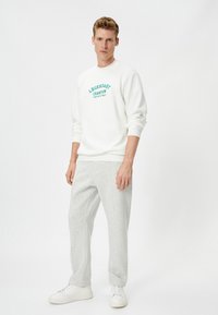Sudadera blanca con letras verdes, combinada con pantalones de chándal grises. Modelo de pie, mostrando un ajuste relajado y mangas acampanadas. Zapatillas blancas.