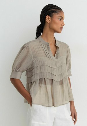 Kvinde med flettet hår iført en beige lagdelt bluse med pufærmer og hvide bukser, stående sidelæns mod en hvid væg.