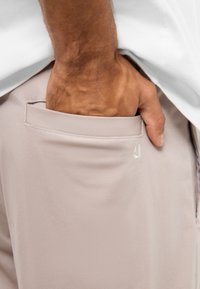 Svetlo béžové nohavice s hladkou textúrou, ktoré majú zadnú kapsu a subtílnu logo detail pri kapsičke.