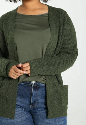 Personne portant un cardigan côtelé vert foncé sur une chemise vert olive et un jean bleu, les mains jointes près de la taille.