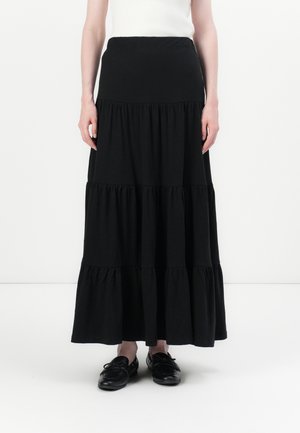 Donna che indossa una lunga gonna nera a balze, top bianco senza maniche e mocassini neri in pelle, in piedi contro uno sfondo bianco.