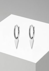 Orecchini a cerchio in argento con un pendente triangolare. Texture liscia e lucidata con una superficie riflettente, attaccati da un piccolo cardine.