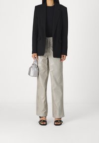 Nainen, jolla on päällään musta bleiseri, krokotiilikuosiset vaaleanharmaat housut, mustat sandaalit, ja hän pitää kädessään pientä hopeista käsilaukkua valkoista taustaa vasten.