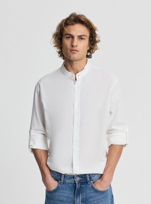 Młody mężczyzna o kręconych włosach, ubrany w białą koszulę ze stójką i podwiniętymi rękawami oraz niebieskie dżinsy, ręce w kieszeniach, neutralne tło.