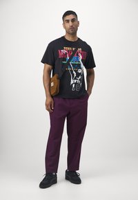 Zwarte graphic t-shirt met kleurrijke print; paarse broek; bruine crossbody tas; zwarte sneakers. Eenvoudig, casual ontwerp met gedurfde kleuren.