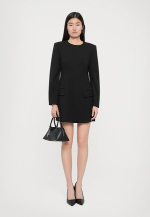 Black shift dress with long sleeves and two front pockets; paired with a small black handbag and pointed black heels. Smooth texture.