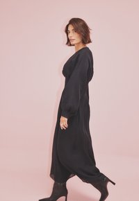 Juoda ilga rankovė su laisvu siluetu ir pūkuotomis rankovėmis, derinama su juodomis iki kelių stiletto batais prieš rožinį foną.