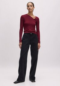 Burgunderfarbenes Longsleeve-Oberteil mit asymmetrischem Ausschnitt, kombiniert mit hochgeschnittenen, schwarzen Weitbeinjeans im klassischen Fünf-Taschen-Design.