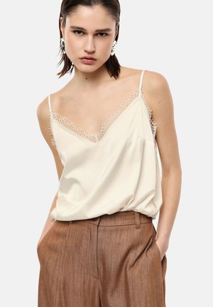 Giovane donna con capelli corti e scuri indossa una canottiera color crema bordata di pizzo e pantaloni marroni a vita alta, mani nelle tasche, sfondo neutro.