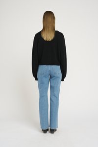 Suéter preto de malha com ombros caídos combinado com jeans azuis de cintura alta. Botas pretas de salto completam o look. O fundo é branco liso.