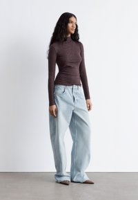 Braun gemusterter Langarm-Oberteil mit figurbetontem Schnitt, kombiniert mit hellblauen, locker sitzenden Jeans und spitzen Schuhen. Glatte Texturen und bequemer Schnitt.