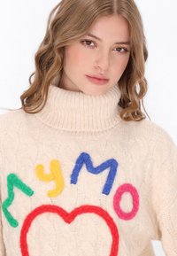 Cremfarbener Strickpullover mit hohem Kragen, der vorne bunte Filzbuchstaben in Grün, Gelb, Blau, Rot und Pink aufweist.