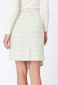 Femme portant une jupe taille haute texturée bleu pastel et crème avec une fente arrière, associée à un haut blanc à manches longues.