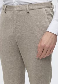 Homme portant un pantalon beige texturé et une chemise blanche boutonnée, la main droite détendue le long du corps.