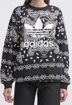 Sweat Adidas noir et blanc à motif cachemire avec grand logo trèfle et nom de la marque centré sur la poitrine, porté par une personne.