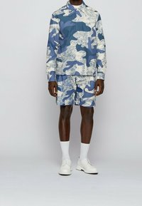 Mies, joka pukeutuu sinivalkoiseen abstraktiin kuviolliseen pitkähihaiseen paitaan ja shortseihin, valkoisiin sukkiin ja valkoisiin nauhakenkiin, seisoo harmaalla lattialla.