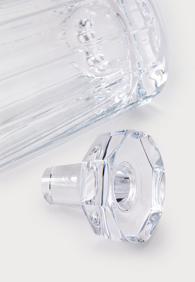 Primer plano de un tapón de vidrio transparente colocado junto al cuello de una decantadora de vidrio acanalado a juego sobre una superficie blanca.