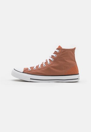 Bruine hoge canvas sneaker met witte veters, witte rubberen zool, zwarte strepen en oranje stiksel detail op de hak tegen een witte achtergrond.