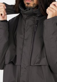 Veste isolante gris foncé avec un col haut, poche avant zippée et texture douce. Présente un logo discret sur la poitrine et des éléments adjustables.