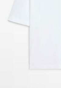 T-shirt blanc à manches courtes suspendu sur un fond blanc, montrant la manche gauche et le bas du côté droit du t-shirt.