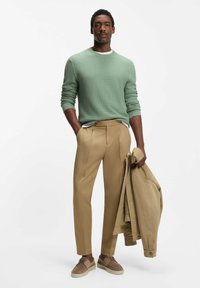 Vihreän mintun sävyinen teksturoitu neule, vaaleanruskeat räätälöidyt plisseehousut ja ruskeat kengät. Beige takki roikkuu hänen käsivarrellaan. Minimalistinen design.